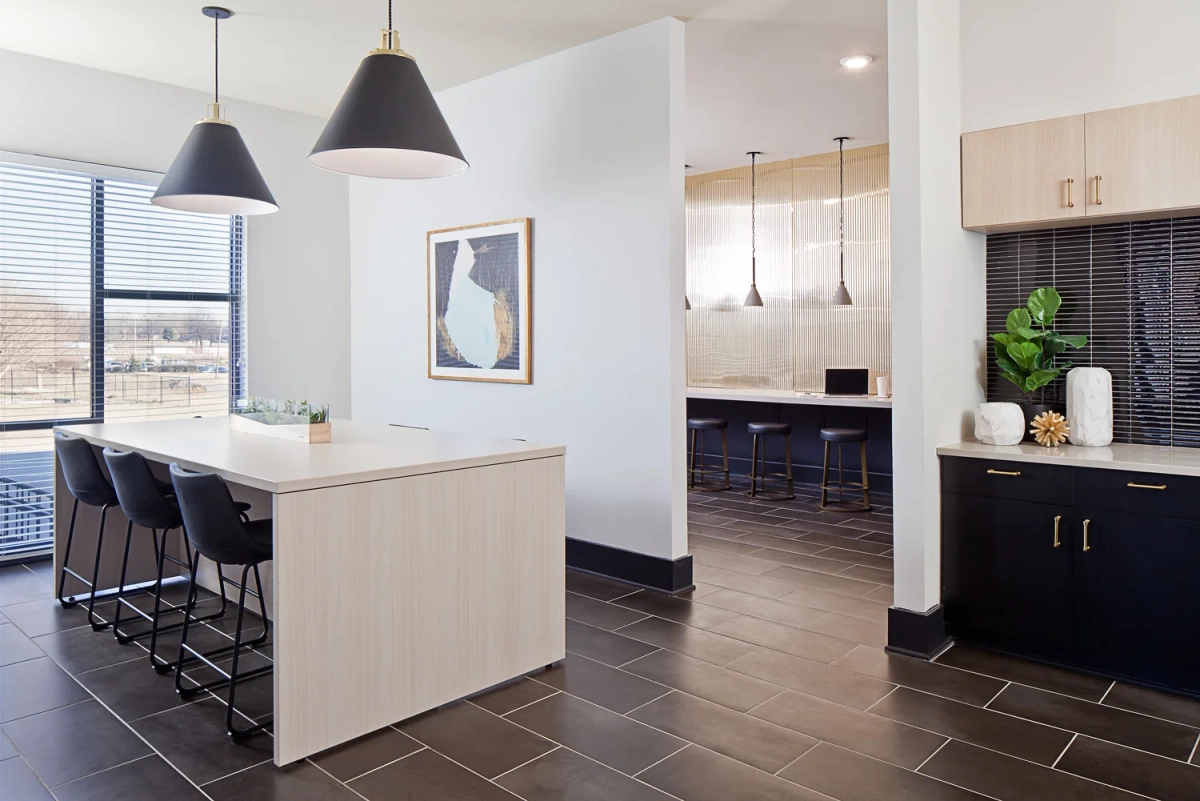 Modern clubhouse kitchen and bar area with white countertop island, black leather bar stools, matte black cone pendant lights, and dark tile backsplash cabinetry with brass hardware