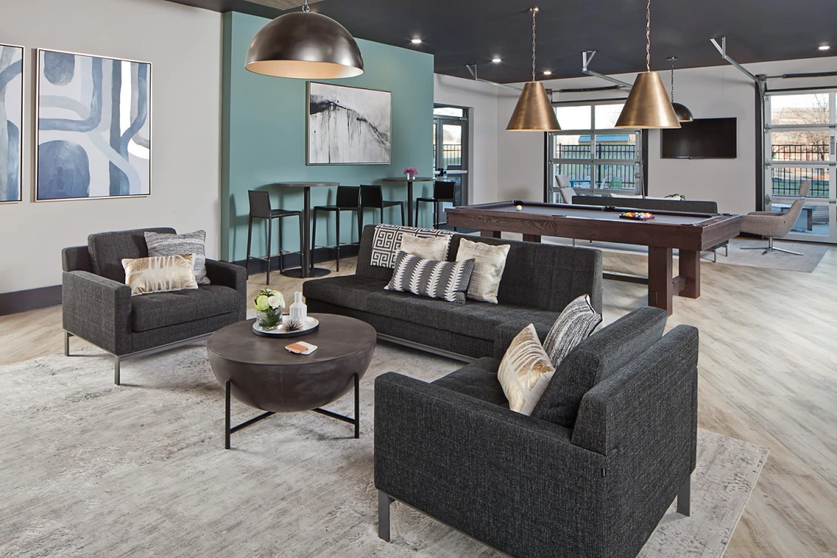 Clubhouse lounge seating area featuring charcoal gray sofa and armchairs with patterned throw pillows, round industrial coffee table, pool table with bronze pendants, and teal accent wall with abstract art