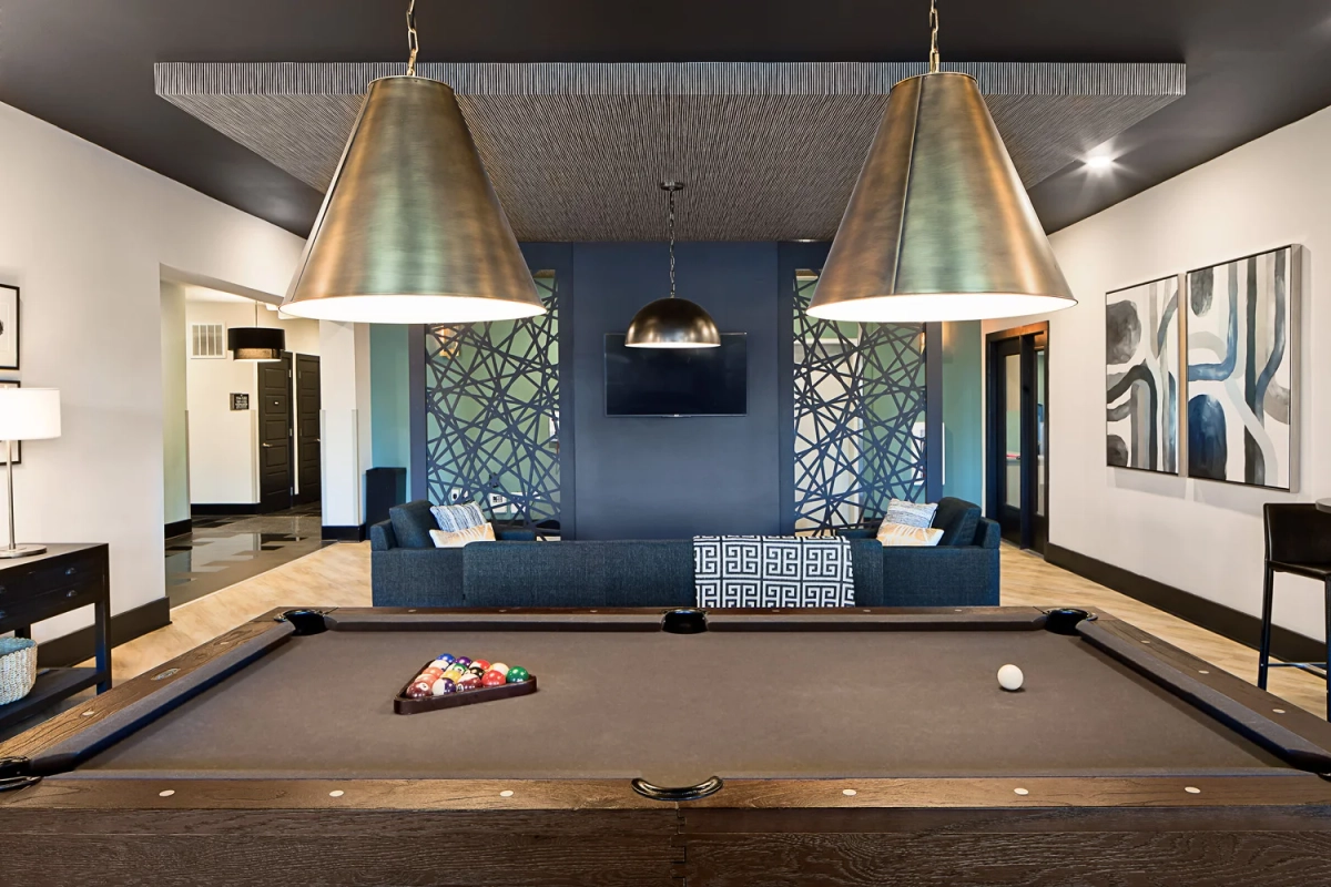 Pool table with racked billiard balls and oversized bronze cone pendant lights, navy blue sectional sofa beyond a geometric metal decorative screen against a dark accent wall