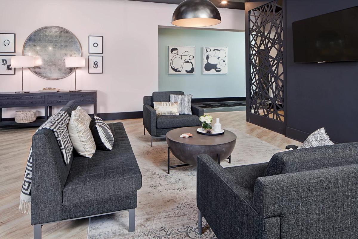 Clubhouse lounge viewed from seating area showing charcoal sofas with geometric pillows, round coffee table, console with antiqued mirror, and decorative metal room divider screen