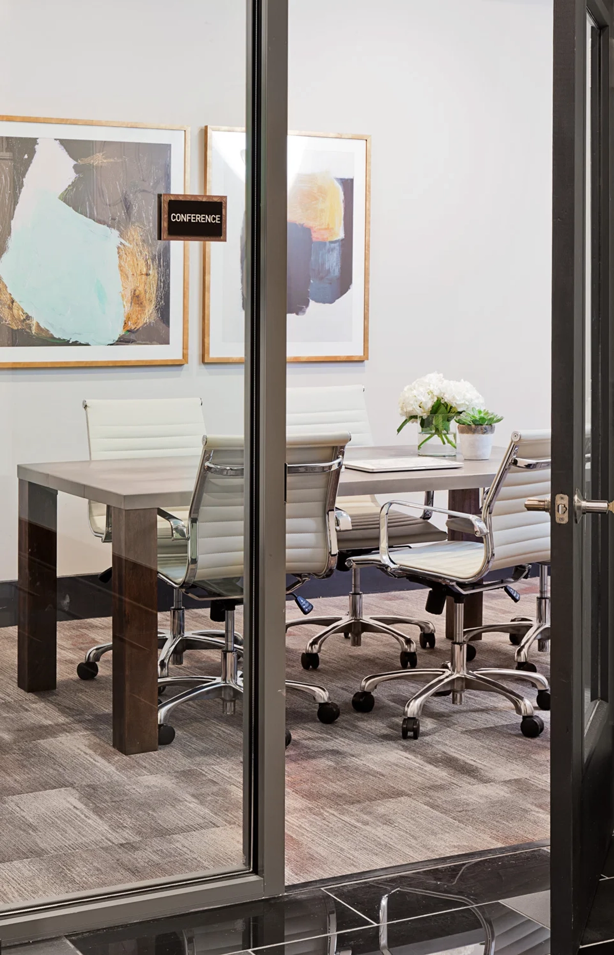 Conference room viewed through glass doors with a gray rectangular table, white leather rolling chairs, abstract artwork, and fresh white hydrangeas