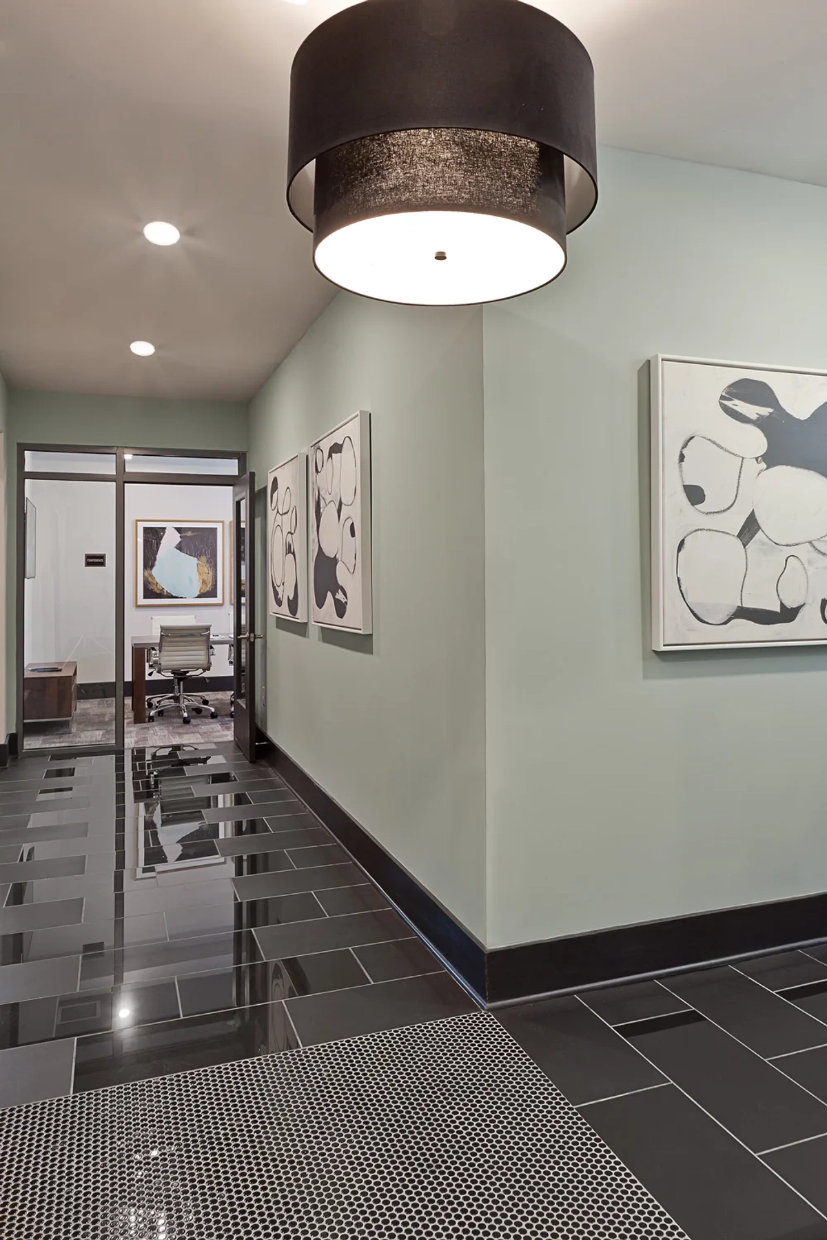 Hallway with polished dark tile flooring and penny-round tile accent strip, sage green walls, large drum ceiling light, and black-and-white abstract artwork leading toward the conference room