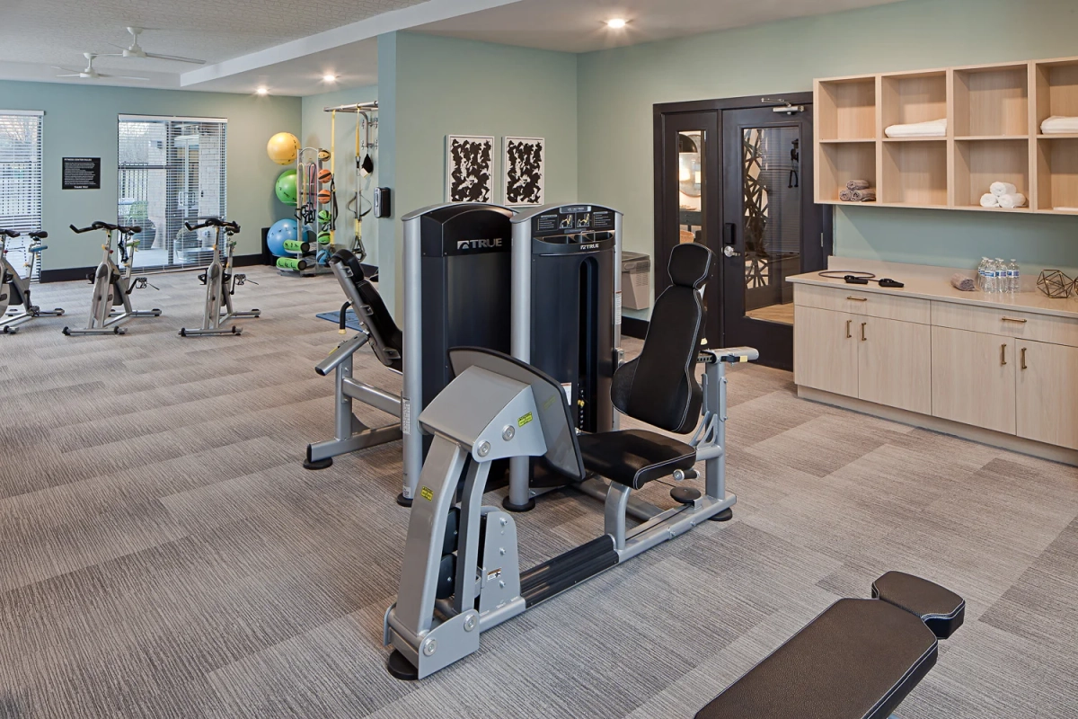 Fitness center with weight machines, spin bikes, exercise balls, sage green accent walls, carpeted flooring, and built-in light wood storage cabinetry with open shelving for towels
