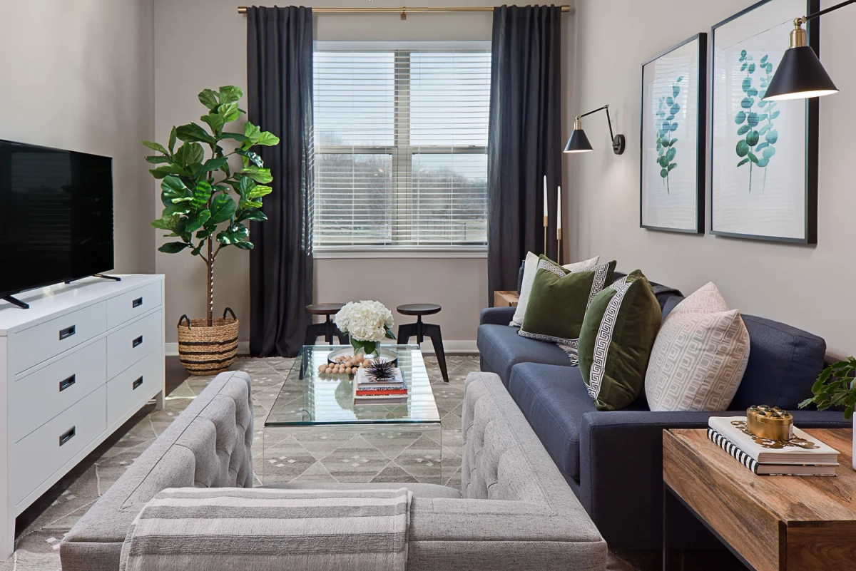 Living room from the TV side showing white media dresser, fiddle leaf fig tree, navy sofa with green pillows, gray armchair, glass coffee table, and navy curtains framing a window