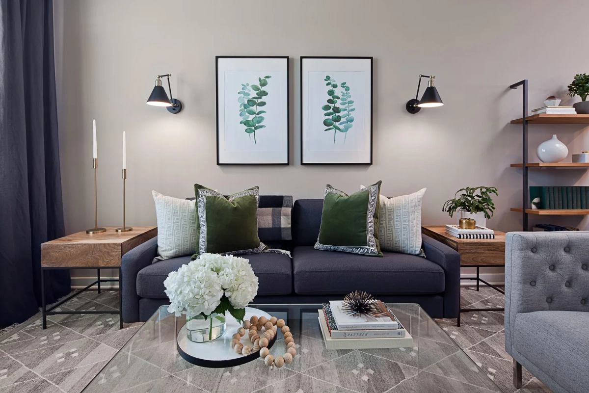 Straight-on view of a navy sofa with green velvet and plaid accent pillows, flanked by black wall sconces, botanical eucalyptus prints above, and glass coffee table with white hydrangeas
