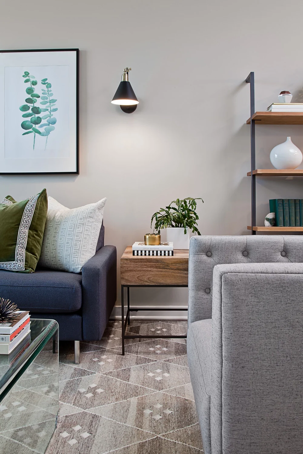 Living room detail showing the corner of a navy sofa, black cone wall sconce, framed eucalyptus print, wood-and-metal side table with potted plant, and gray tufted armchair