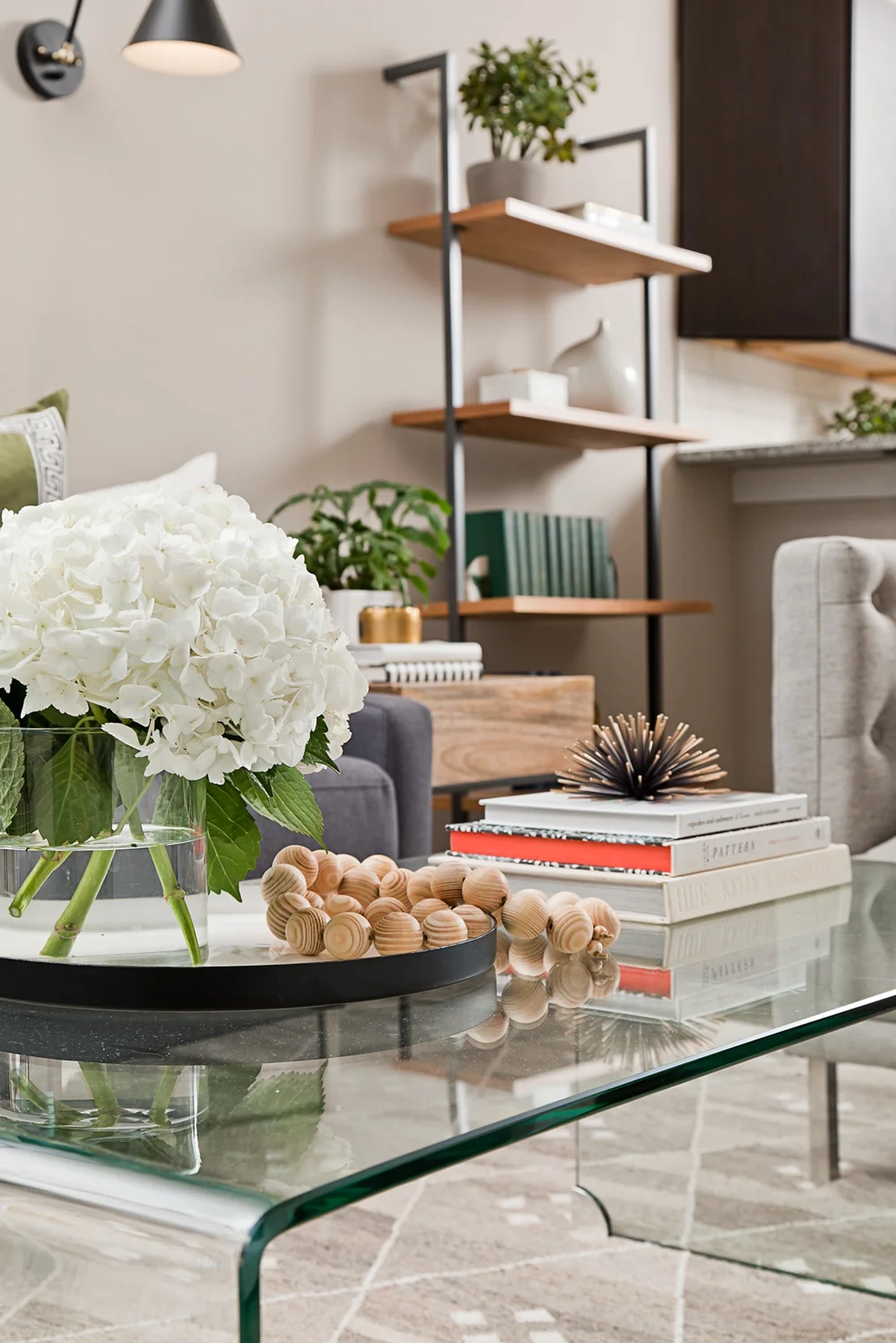 Close-up of a glass coffee table styled with white hydrangeas, wooden bead garland, stacked design books, and a spiky brass sculptural accent