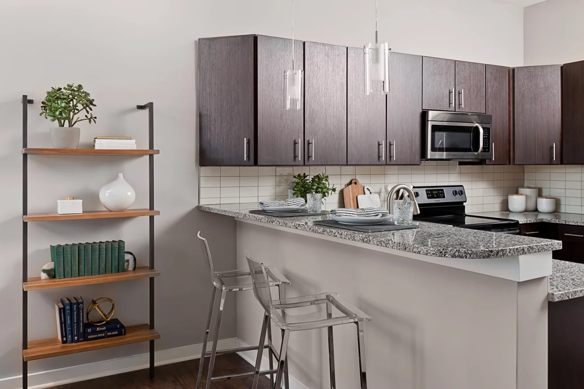 Apartment kitchen with dark espresso upper cabinets, granite countertop peninsula with clear acrylic bar stools, white subway tile backsplash, and stainless steel appliances