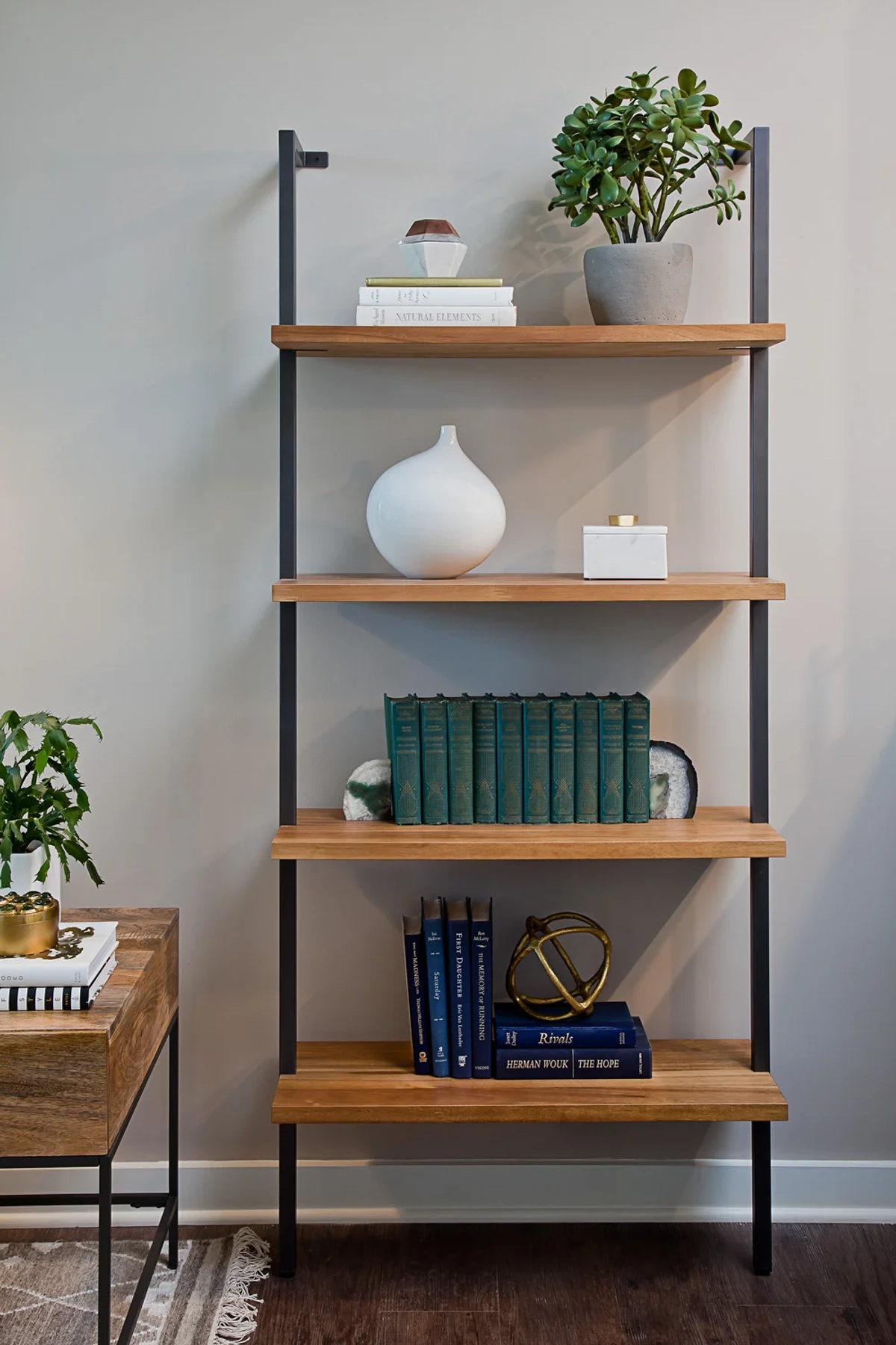Close-up of a wood-and-metal ladder bookshelf styled with a white ceramic vase, green books, brass armillary sphere, and a potted jade plant