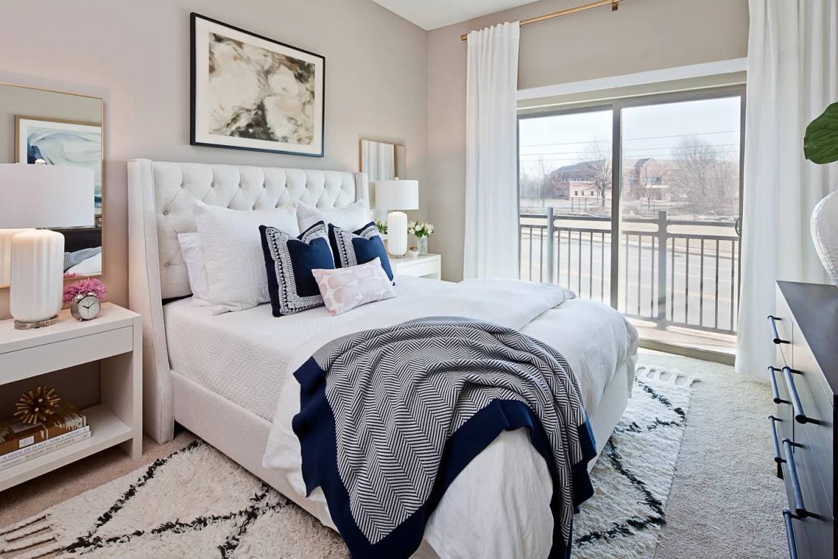 Bright apartment bedroom with white tufted upholstered bed, navy and white embroidered pillows, pink accent pillow, herringbone throw blanket, and dark navy dresser with abstract art