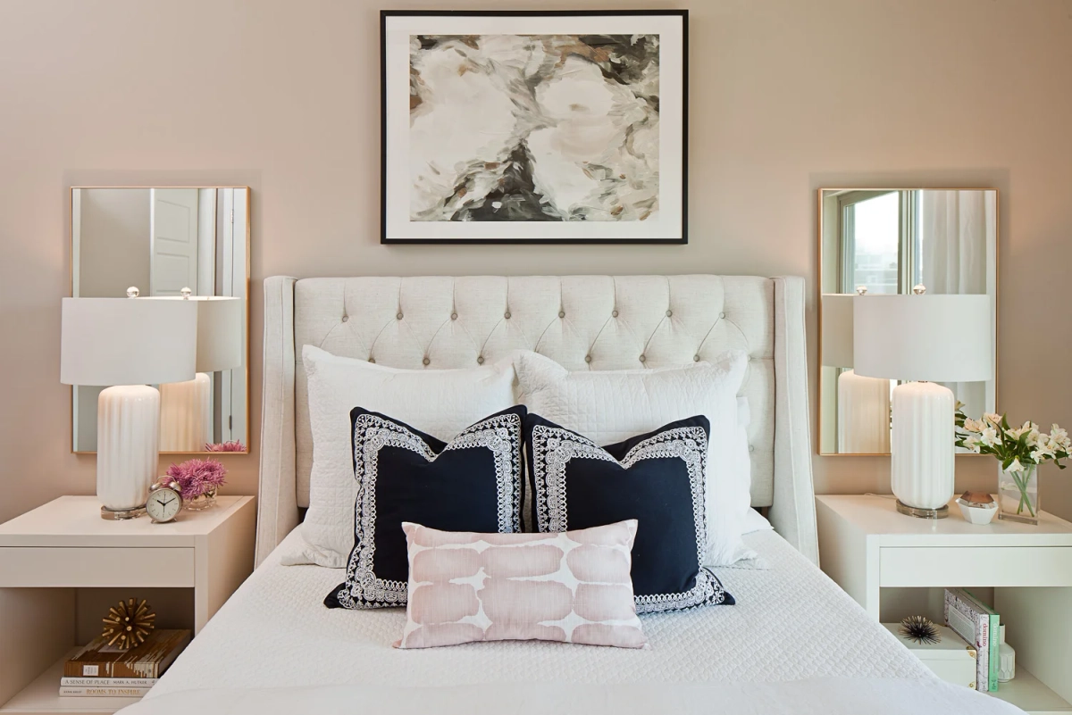 Bedroom headboard detail with white tufted upholstered bed, symmetrical white nightstands with ribbed ceramic lamps, navy embroidered pillows, and abstract marble artwork above