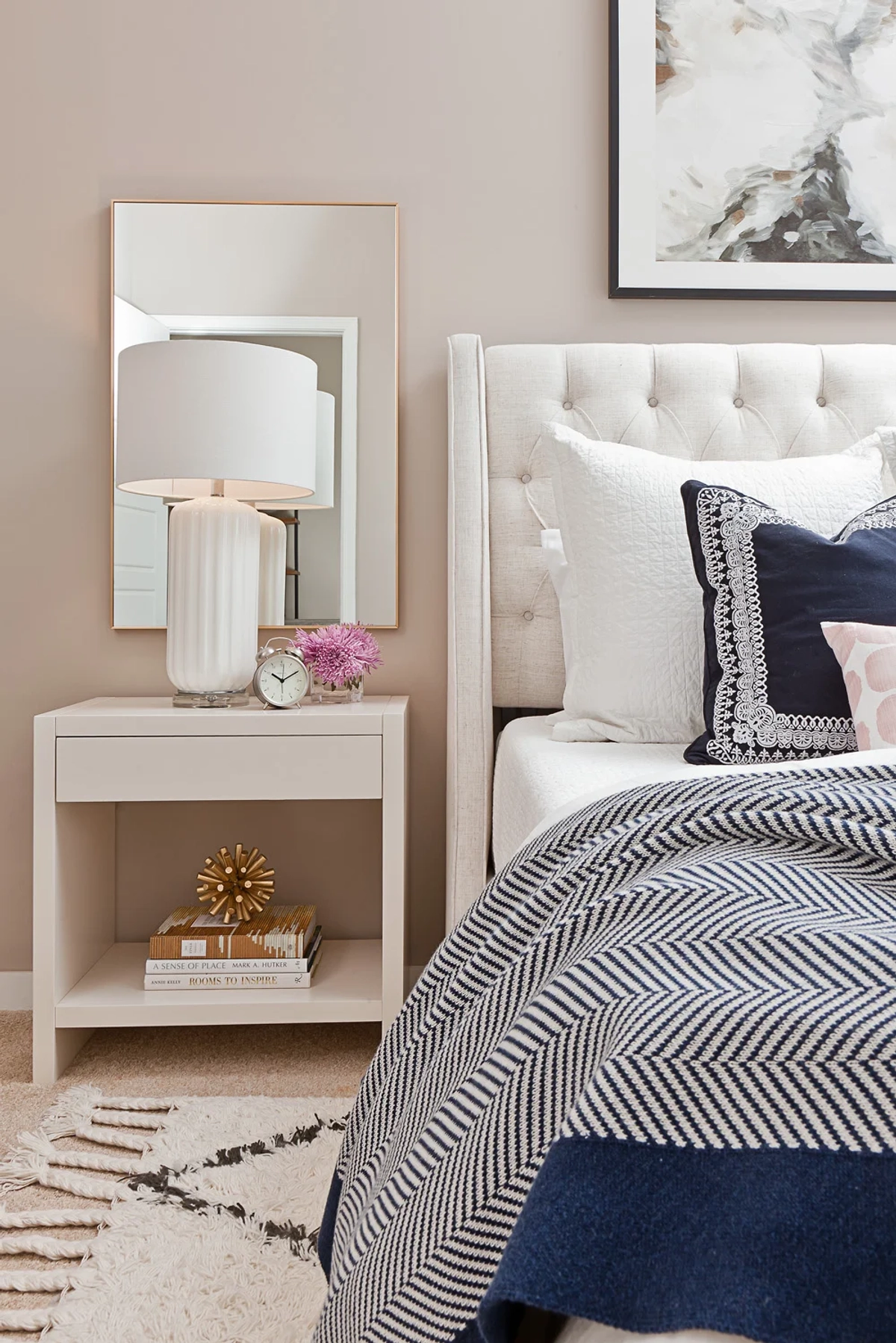 Nightstand detail with white open-shelf nightstand, ribbed white ceramic table lamp, gold-framed mirror, alarm clock, and stacked books on a plush shag rug