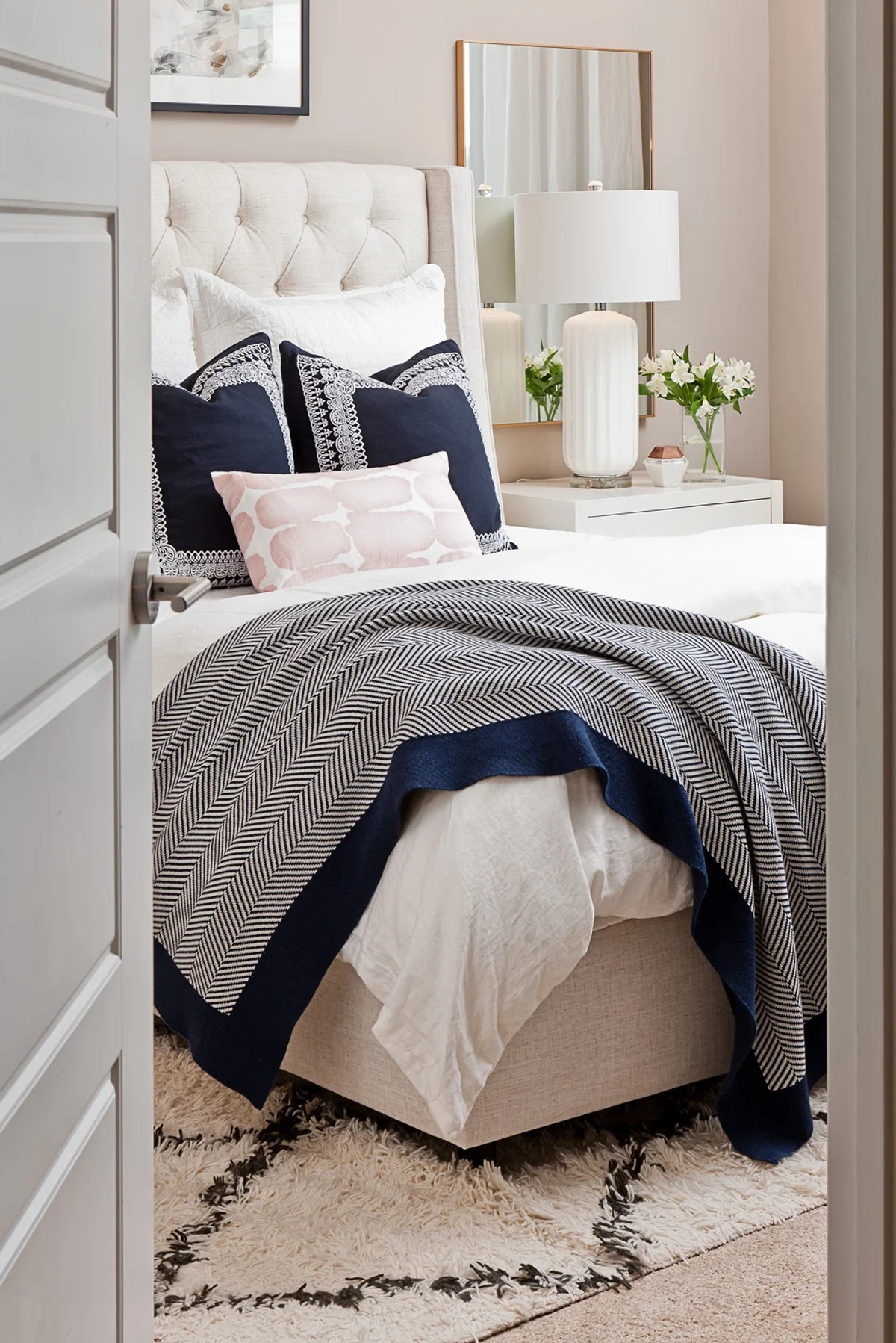 Bedroom viewed through a doorway showing white tufted bed with navy embroidered pillows, herringbone throw blanket, nightstand with lamp and fresh flowers, and black-and-white Moroccan shag rug