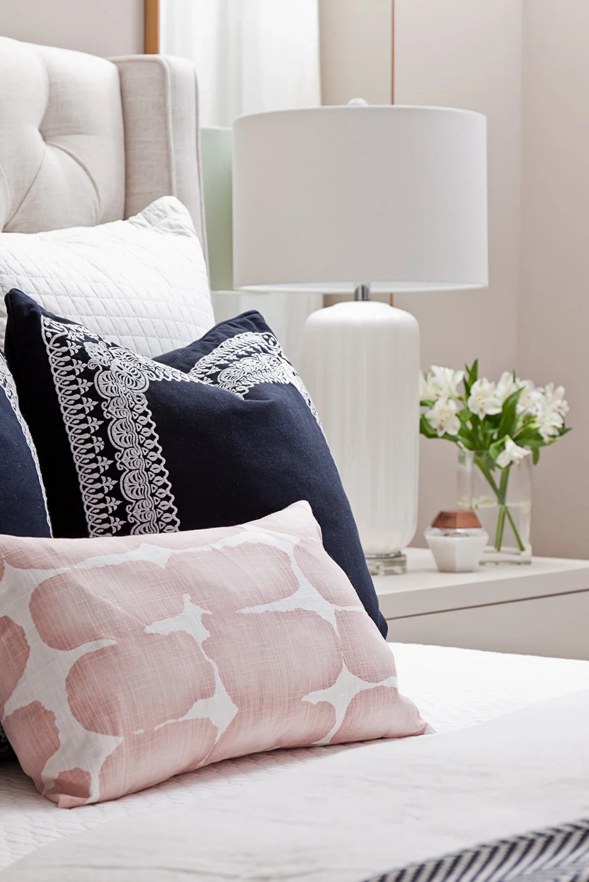 Close-up of decorative bed pillows featuring a pink trellis-patterned lumbar pillow, navy blue embroidered euro sham, white quilted pillow, and a white ceramic lamp with fresh flowers