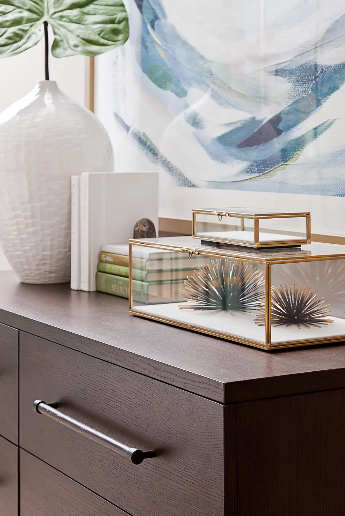 Dresser top styling detail with dark wood dresser, white textured ceramic vase with monstera leaf, brass-and-glass display boxes, stacked books, and a blue-green abstract painting above