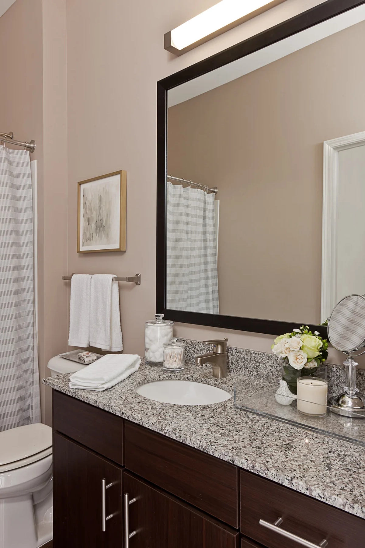 Apartment bathroom with dark espresso vanity, granite countertop, large dark-framed mirror, brushed nickel fixtures, striped shower curtain, and fresh flowers