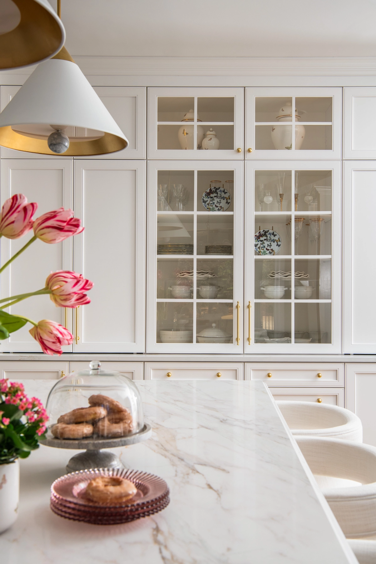 Glass-front kitchen cabinets with curated dishware, brass hardware, and marble countertop with tulips