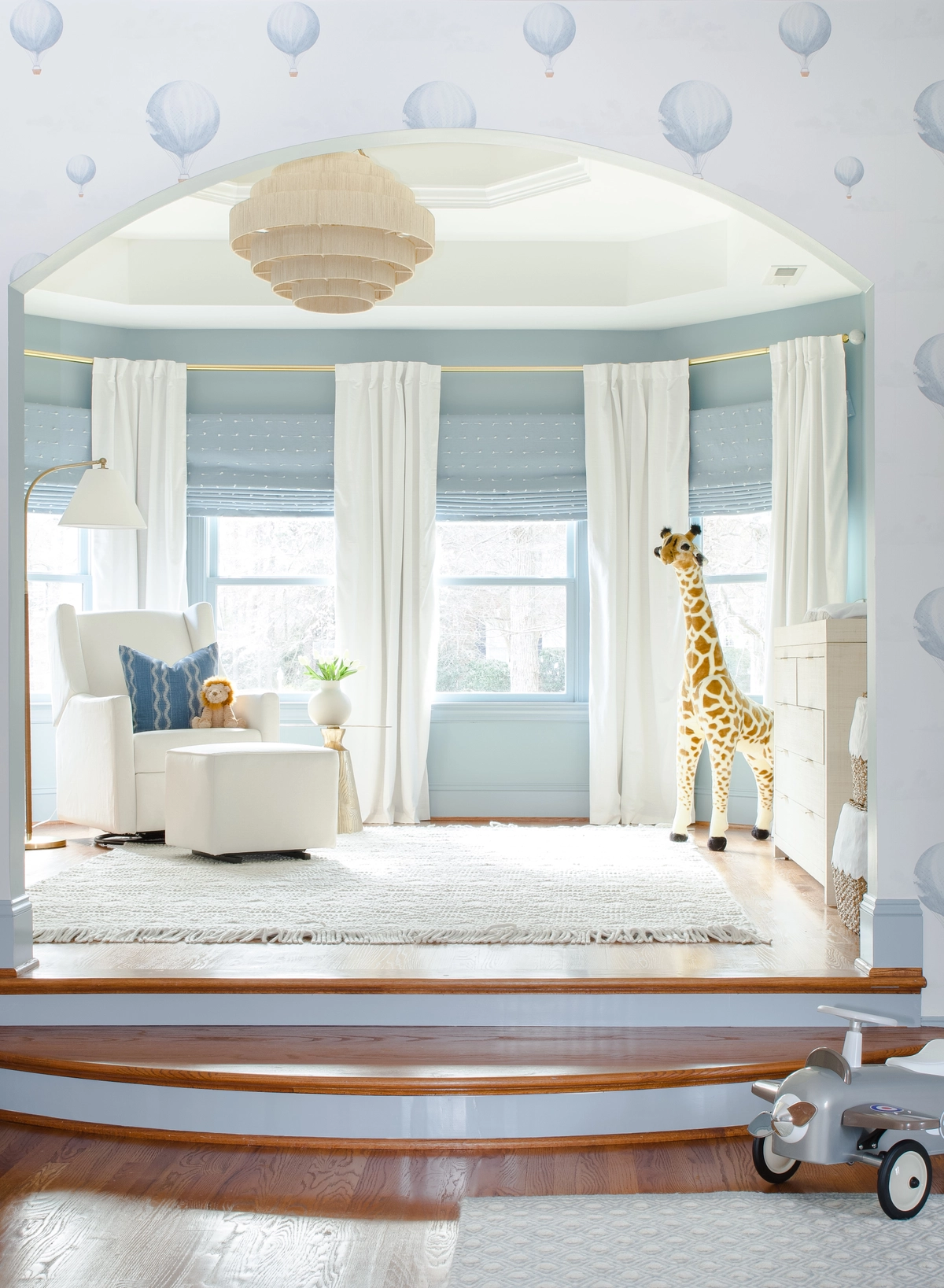 Nursery viewed through an arched doorway with hot air balloon wallpaper, white glider chair, stuffed giraffe, white dresser, blue roman shades, and a tiered pendant light