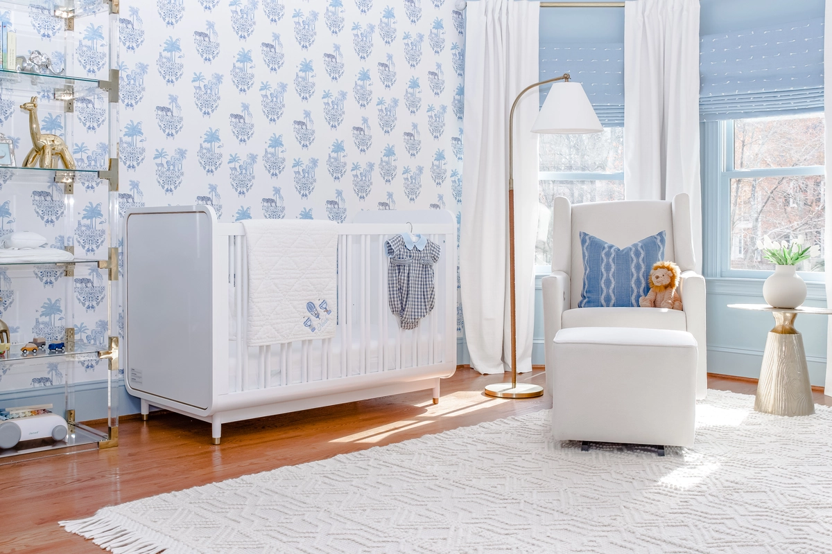 Nursery with blue toile wallpaper, white crib, white glider chair with blue pillow, brass floor lamp, acrylic and brass bookshelf, and blue roman shades