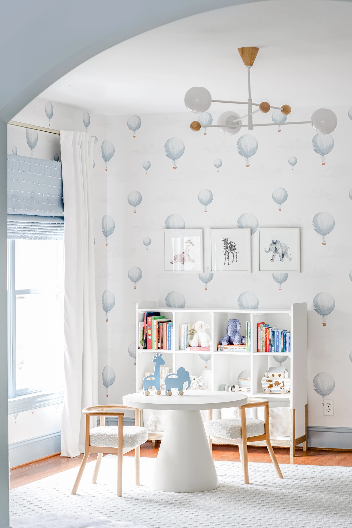 Nursery play nook through an archway with hot air balloon wallpaper, white pedestal table and chairs, cube bookshelf with toys, animal art prints, and a modern sputnik light