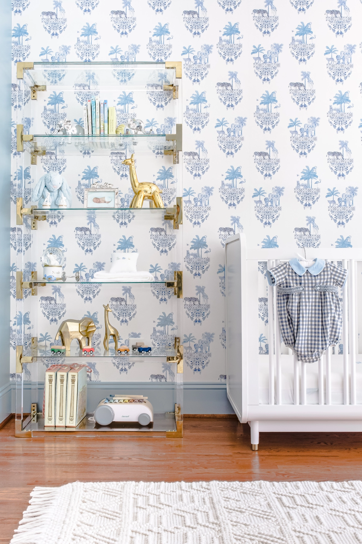 Nursery detail with acrylic and brass bookshelf displaying gold giraffe figurine, toys, and books against blue toile wallpaper, beside a white crib with gingham outfit