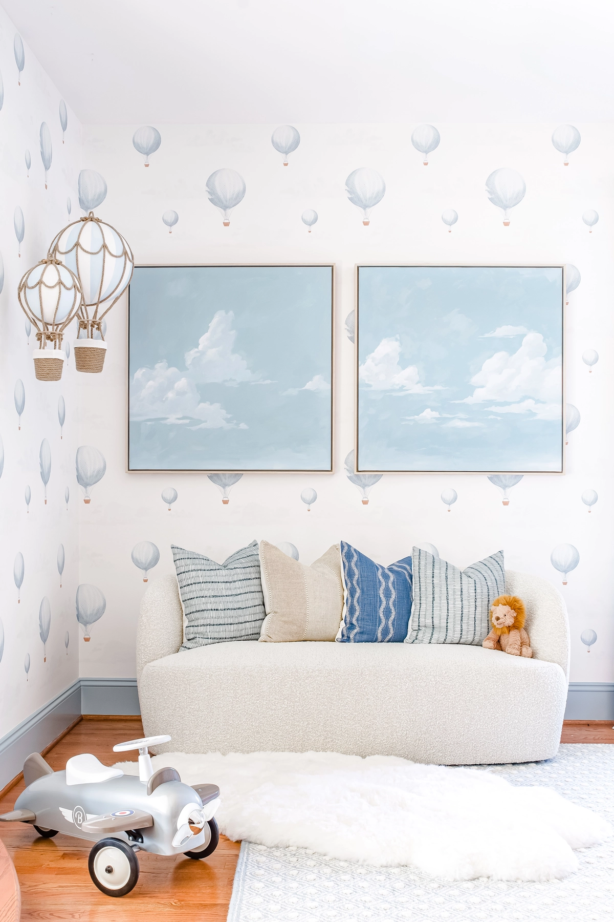 Nursery corner with a cream boucle sofa, blue and neutral accent pillows, paired cloud paintings, hanging hot air balloon mobiles, and hot air balloon wallpaper
