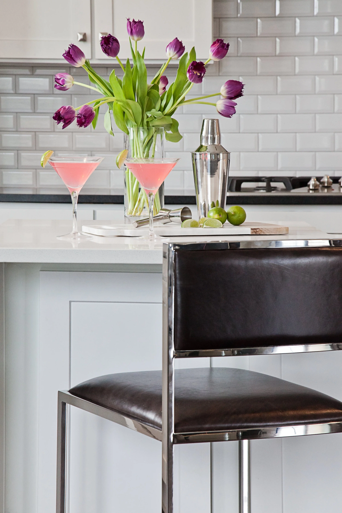 Kitchen island close-up with pink cocktails in martini glasses, silver cocktail shaker, purple tulips in a glass vase, and a brown leather bar stool against white subway tile