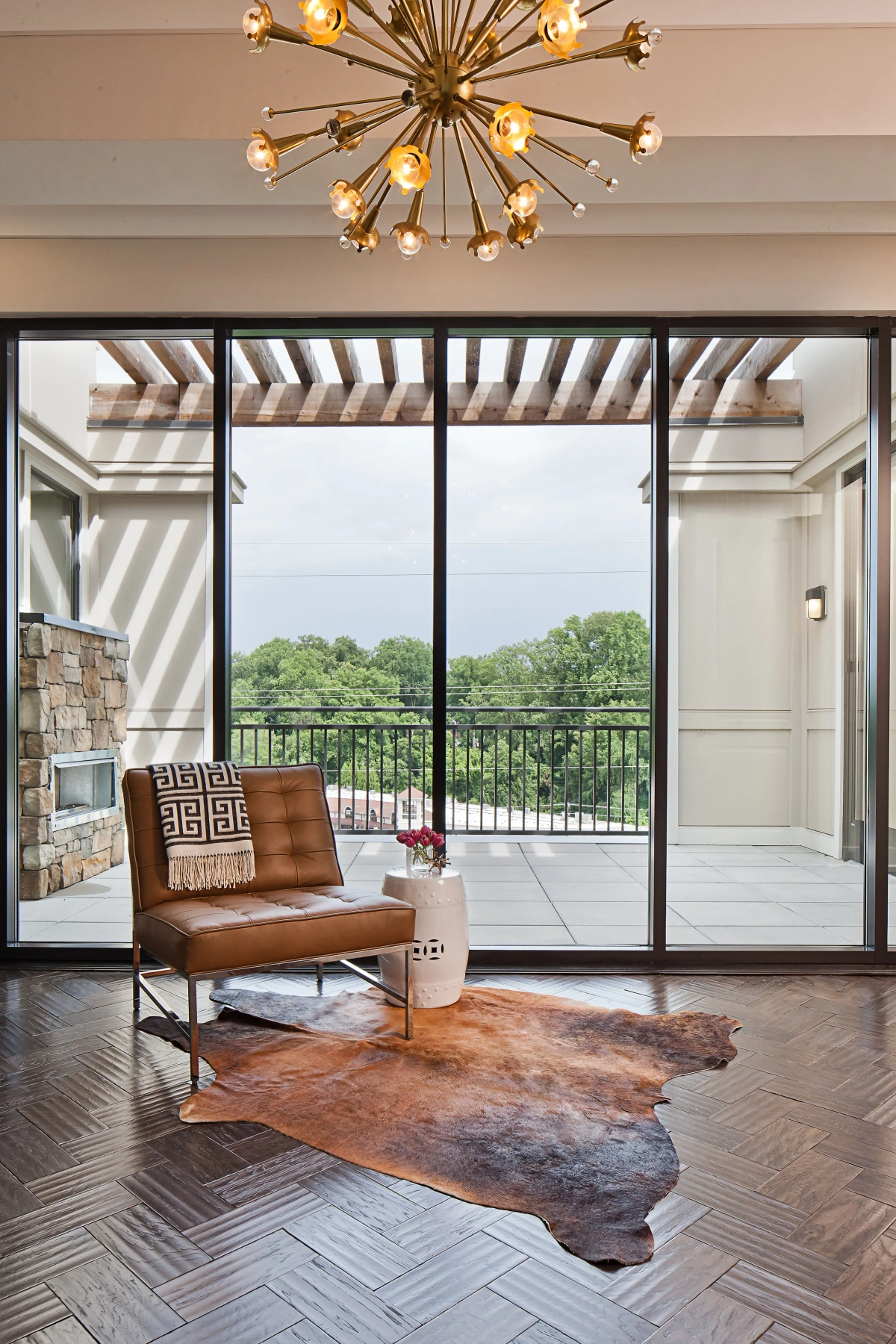 Living area with cognac leather Barcelona-style chair, Greek key throw pillow, white ceramic garden stool, cowhide rug on herringbone wood floors, brass sputnik chandelier, and floor-to-ceiling sliding glass doors