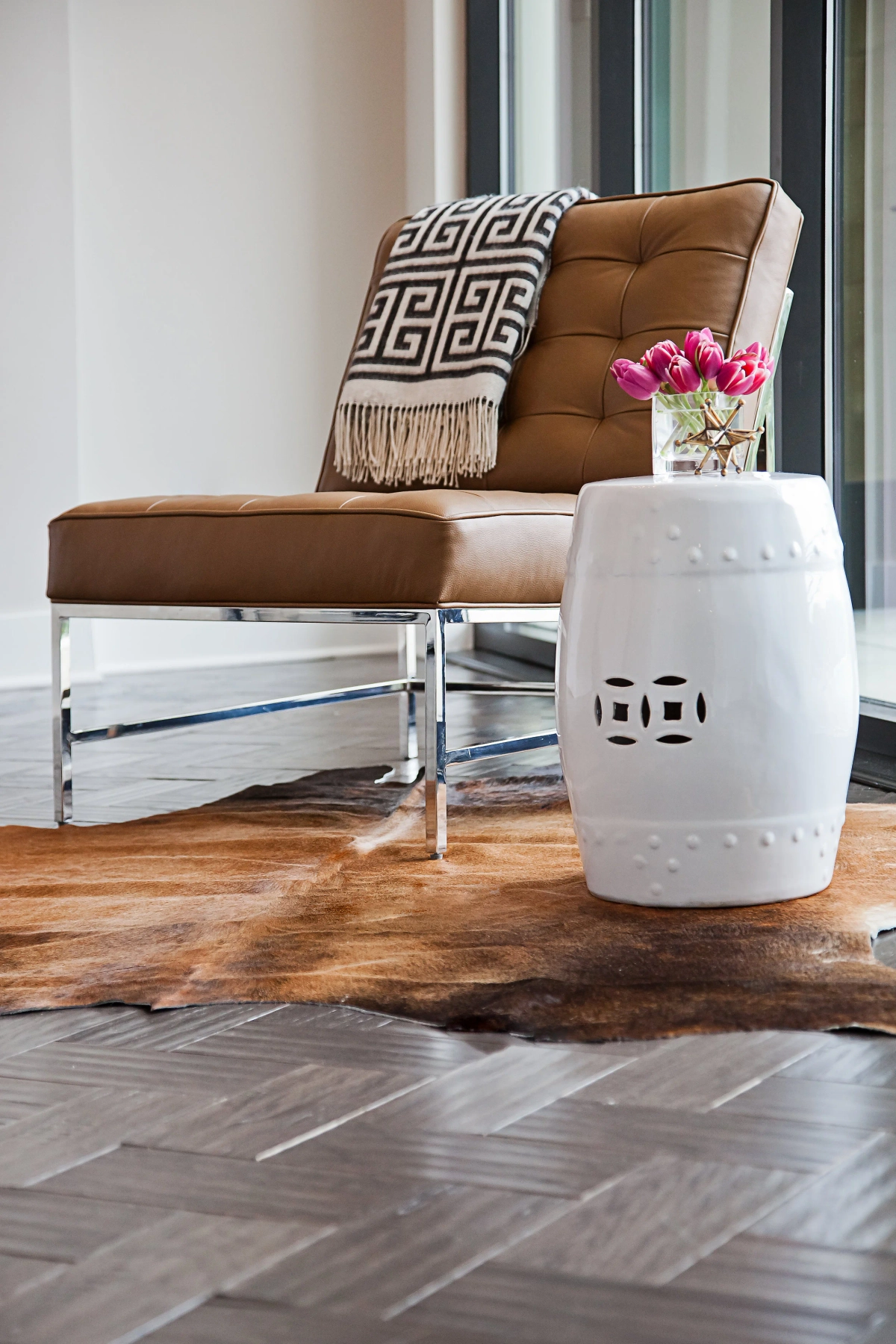 Detail of cognac tufted leather Barcelona chair with geometric black-and-white throw, white ceramic garden stool with pink tulips, cowhide area rug on herringbone hardwood flooring
