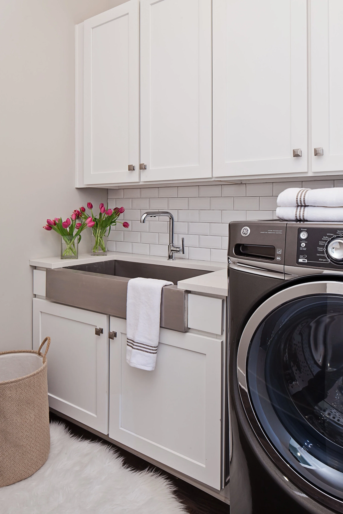Penthouse laundry room with white shaker cabinets, subway tile backsplash, stainless steel farmhouse sink, front-loading washer, sheepskin rug, and woven laundry basket with pink tulips