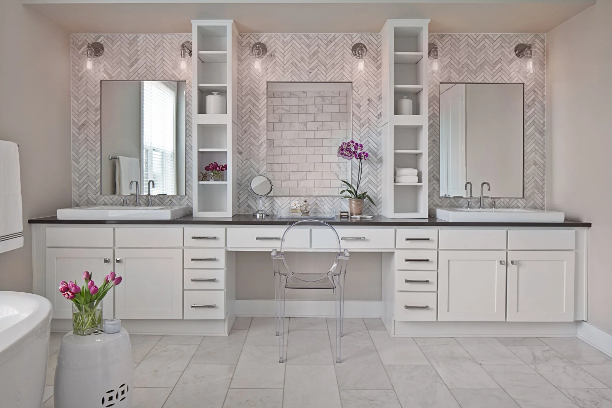 Spacious primary bathroom with white double vanity, herringbone marble tile backsplash, dark countertops, lucite ghost chair at a makeup vanity, open shelving with orchid, and marble tile flooring