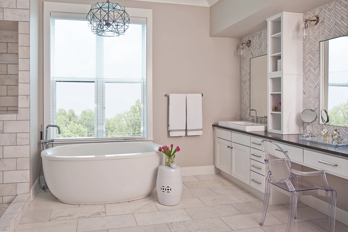 Primary bathroom wide view with freestanding white soaking tub by a large window, geometric orb chandelier, herringbone marble tile accent wall, white vanity with dark countertop, and lucite ghost chair