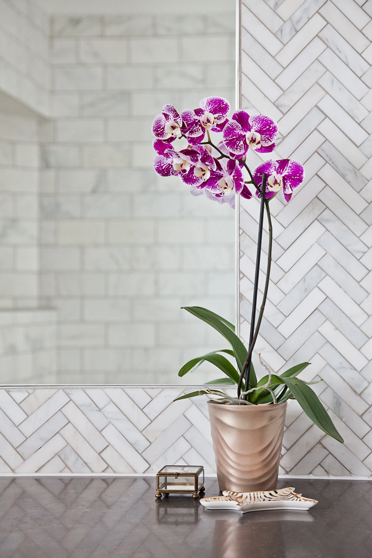Bathroom vanity detail with purple orchid in a blush ceramic pot, small brass jewelry box, and decorative dish against herringbone marble tile and a mirror reflecting the walk-in shower