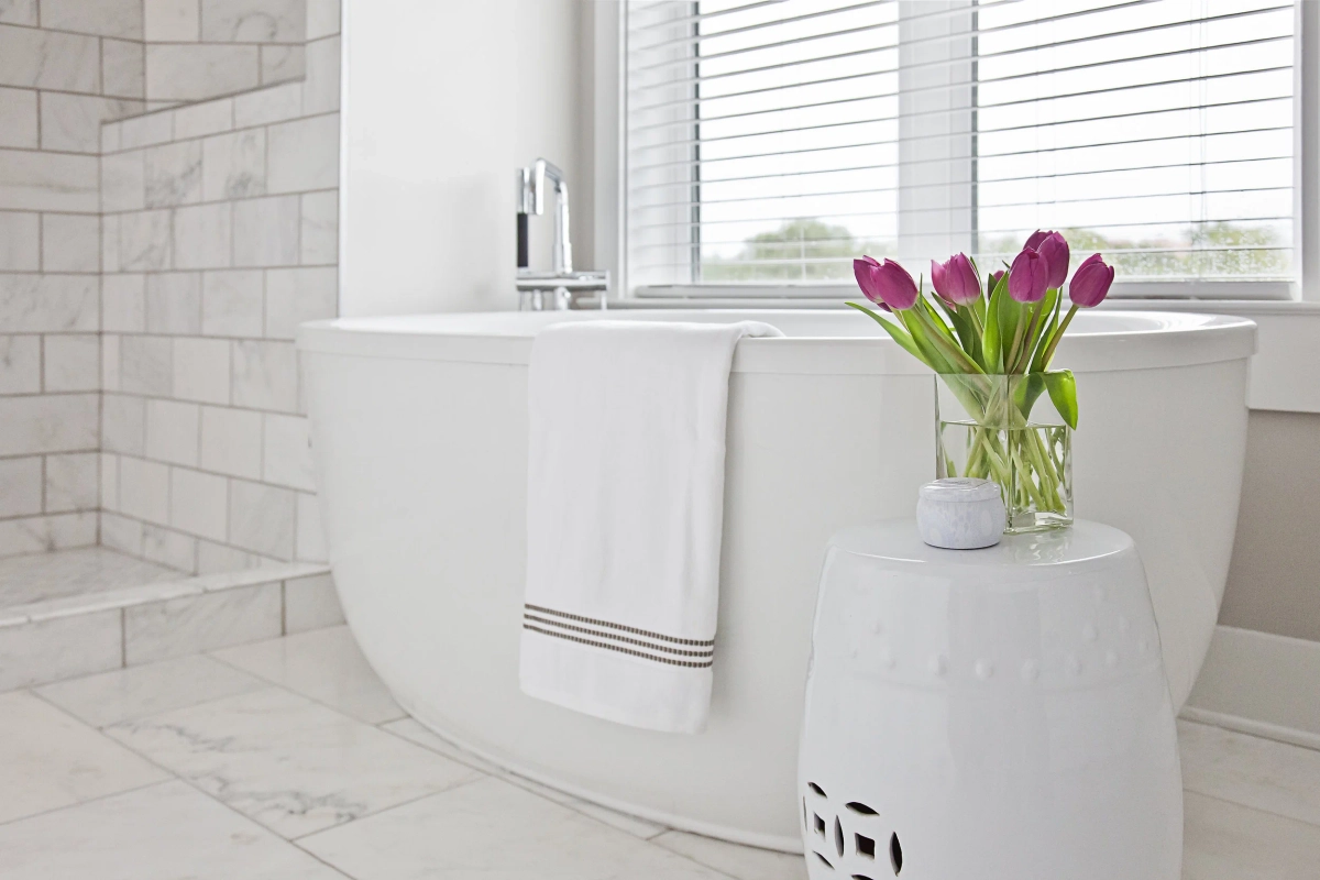 Freestanding white soaking tub detail with folded white towels, purple tulips in a glass vase on a white ceramic garden stool, marble tile floor, and natural light from a window