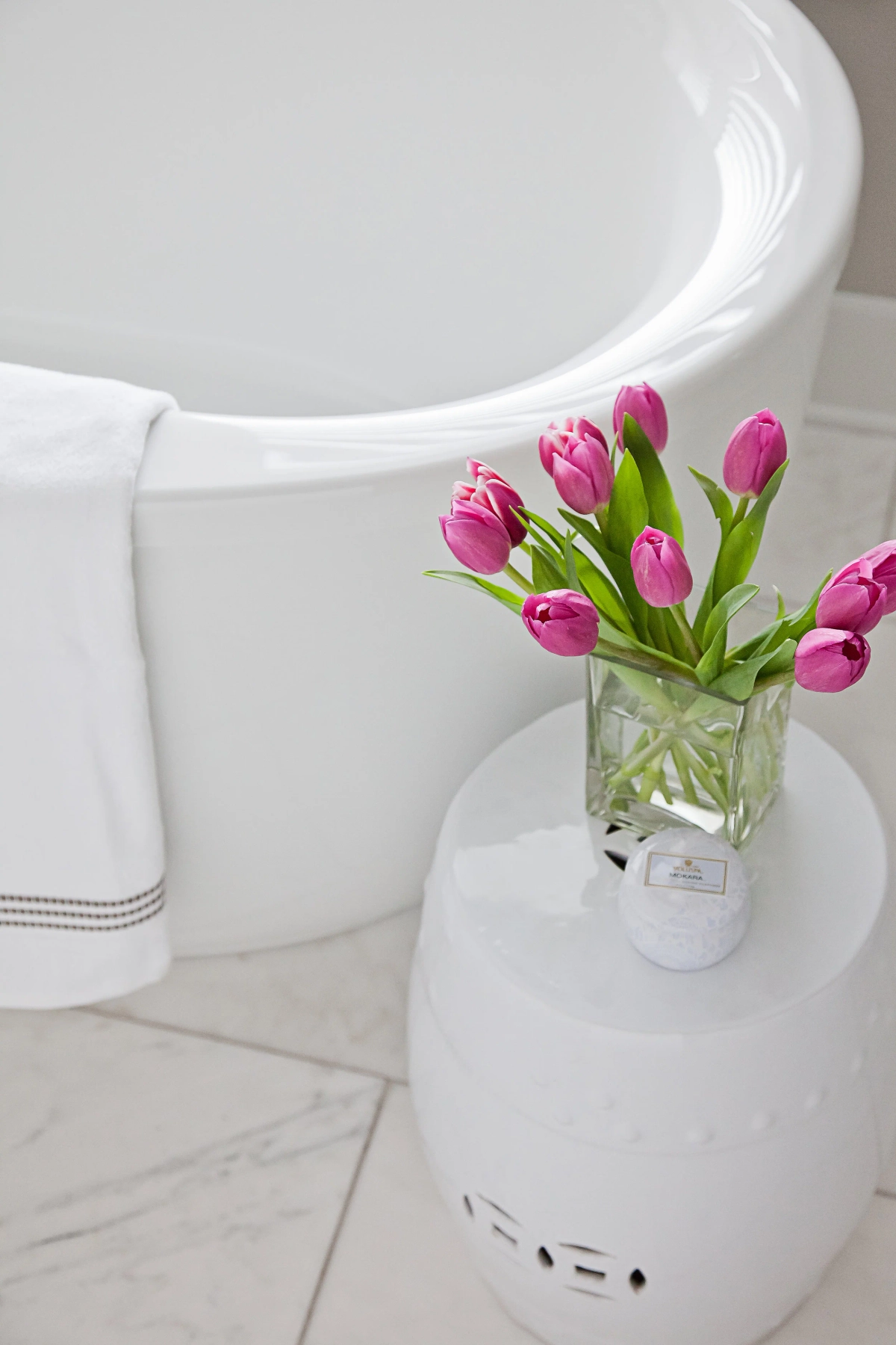 Close-up of a white freestanding tub with pink tulips in a square glass vase on a white ceramic garden stool, white towel with striped trim, and Carrara marble tile flooring