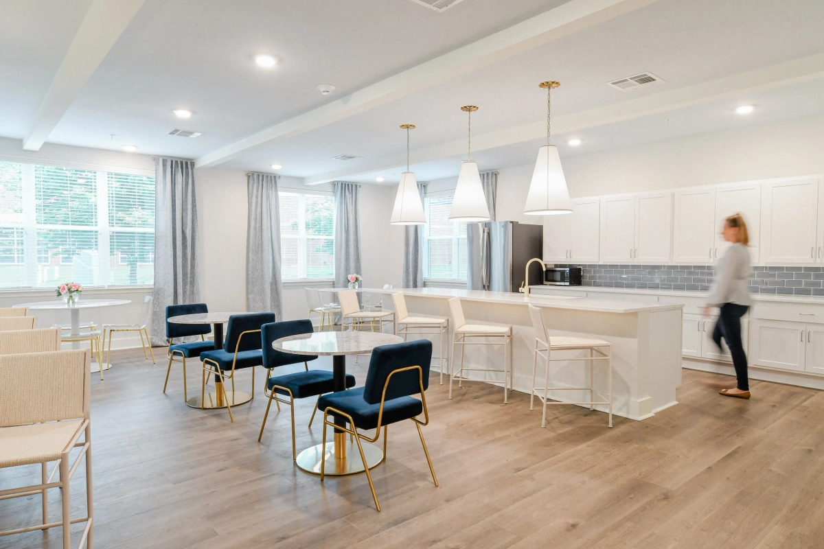 Open-plan senior community dining and kitchen area with round white tables, navy and gold dining chairs, white cone pendant lights over a bar counter, gray subway tile backsplash, and white cabinetry
