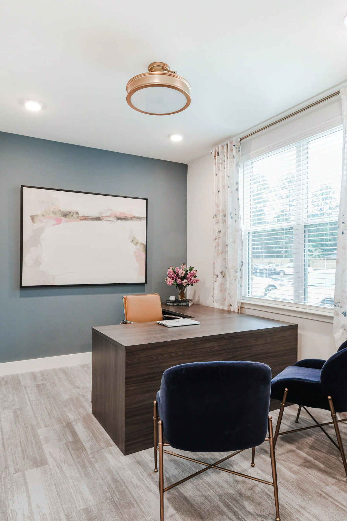 Private office with dark blue-gray accent wall, walnut executive desk, navy velvet guest chairs with gold legs, abstract landscape painting, rose gold flush-mount ceiling light, and floral drapery