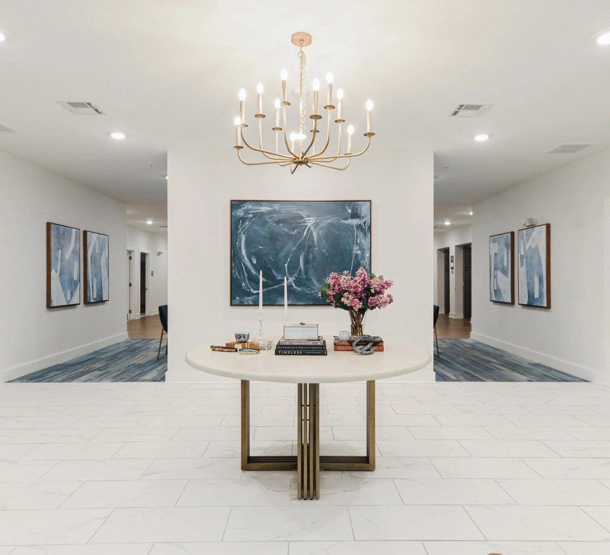 Grand community lobby with white tile floors, round entry table with brass base, pink flower arrangement, gold candelabra chandelier, and a large blue abstract painting