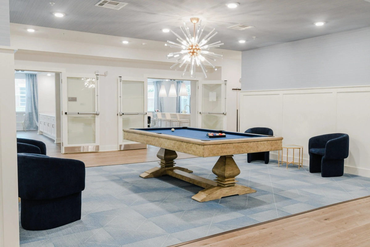 Community game room with a rustic wood pedestal pool table on blue felt, sputnik crystal chandelier, navy swivel club chairs, wainscoting, and glass barn doors opening to the bar area