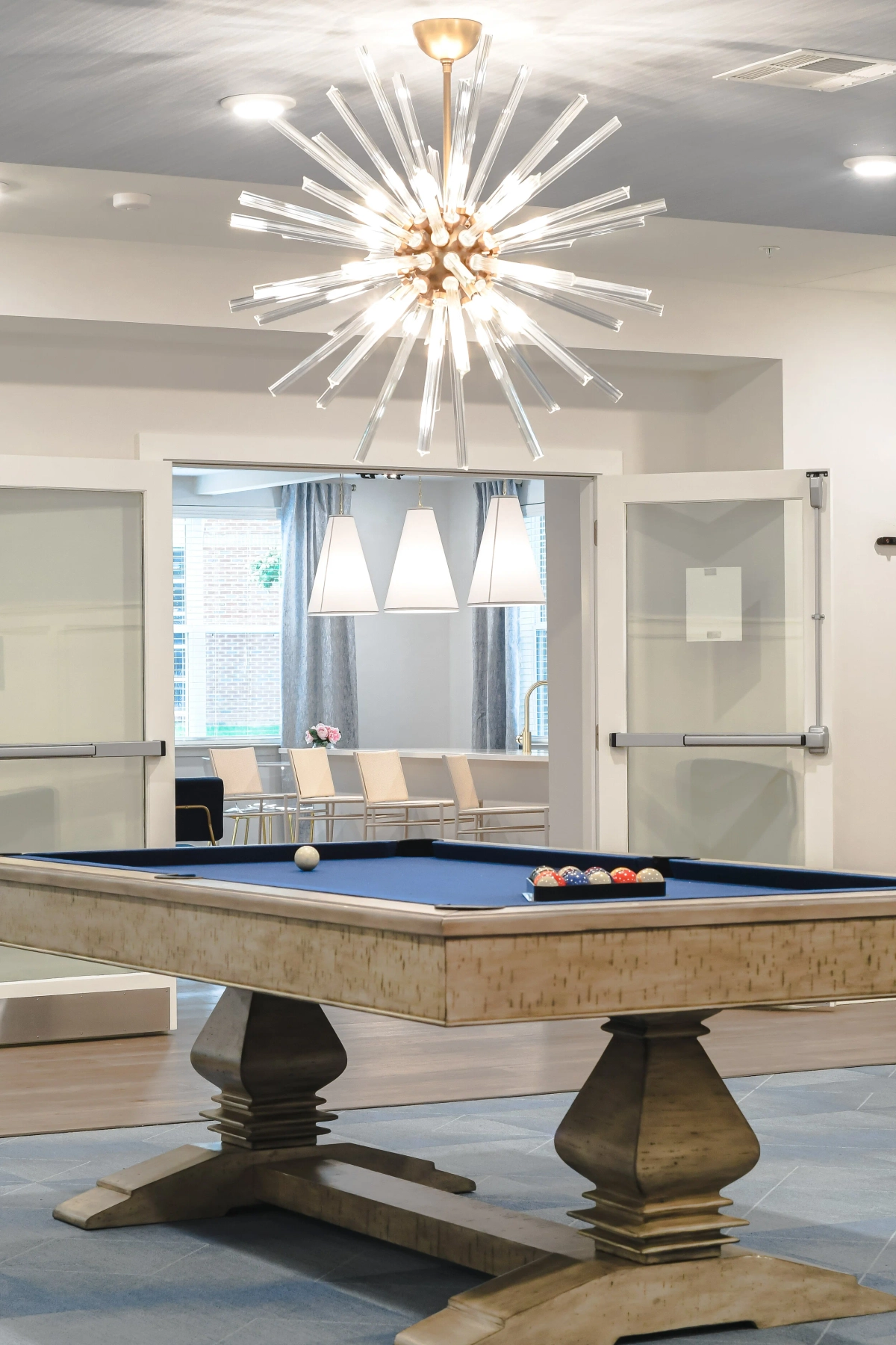 Pool table close-up showing the rustic carved wood pedestal base, blue felt surface with racked balls, dramatic sputnik crystal chandelier overhead, and glass barn doors with white pendant lights