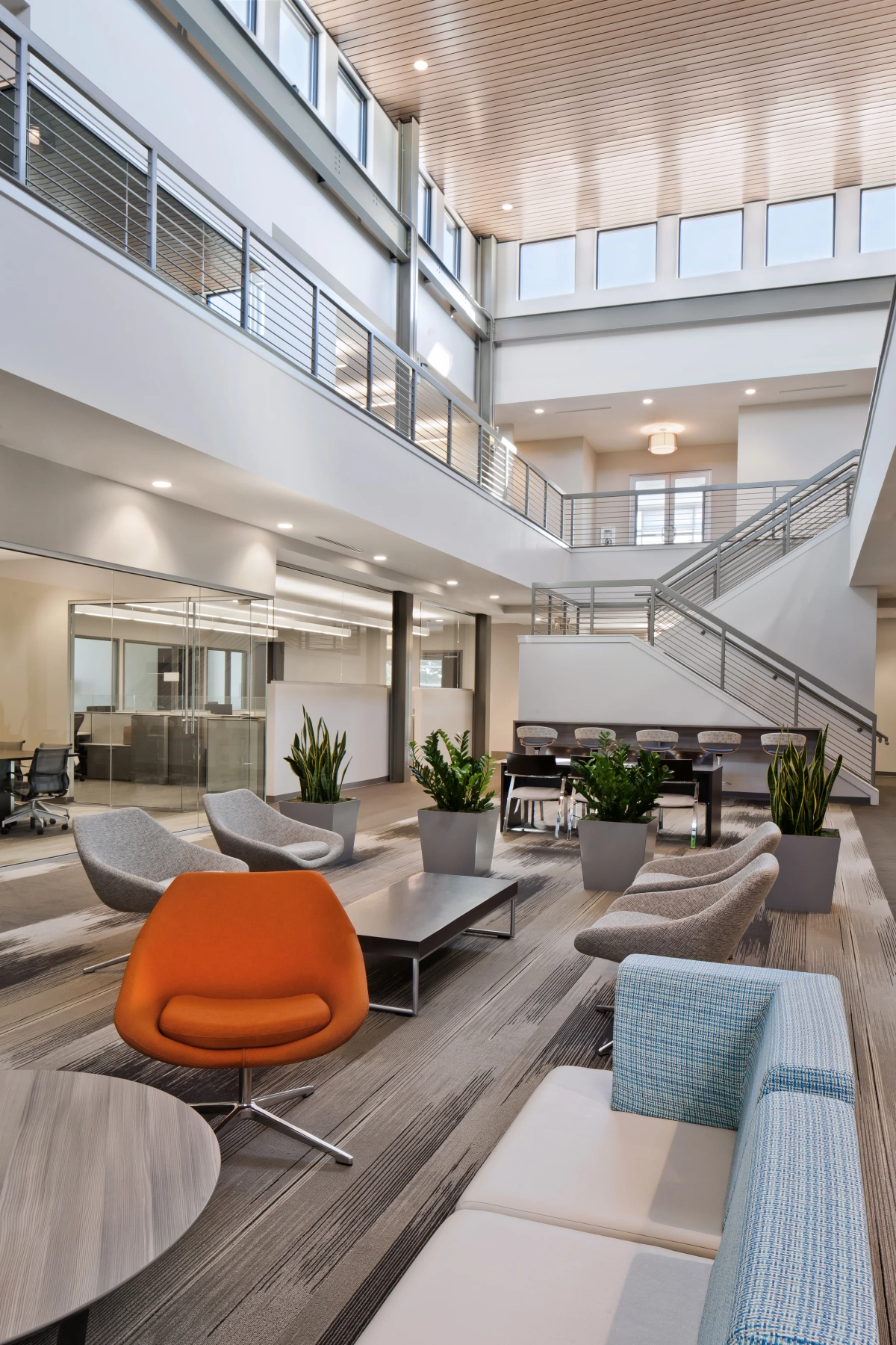 Two-story corporate atrium lounge with orange and gray modern swivel chairs, light blue plaid sofa, potted snake plants, wood-plank flooring, natural wood ceiling accents, and an open staircase