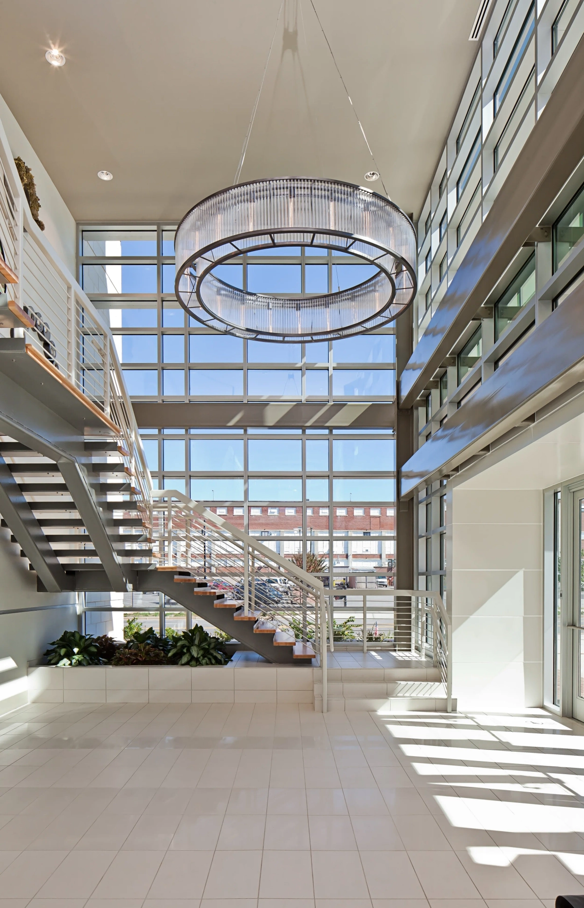 Dramatic double-height office lobby with floor-to-ceiling glass curtain wall, floating open-tread staircase, large oval crystal chandelier, and white tile flooring flooded with natural sunlight