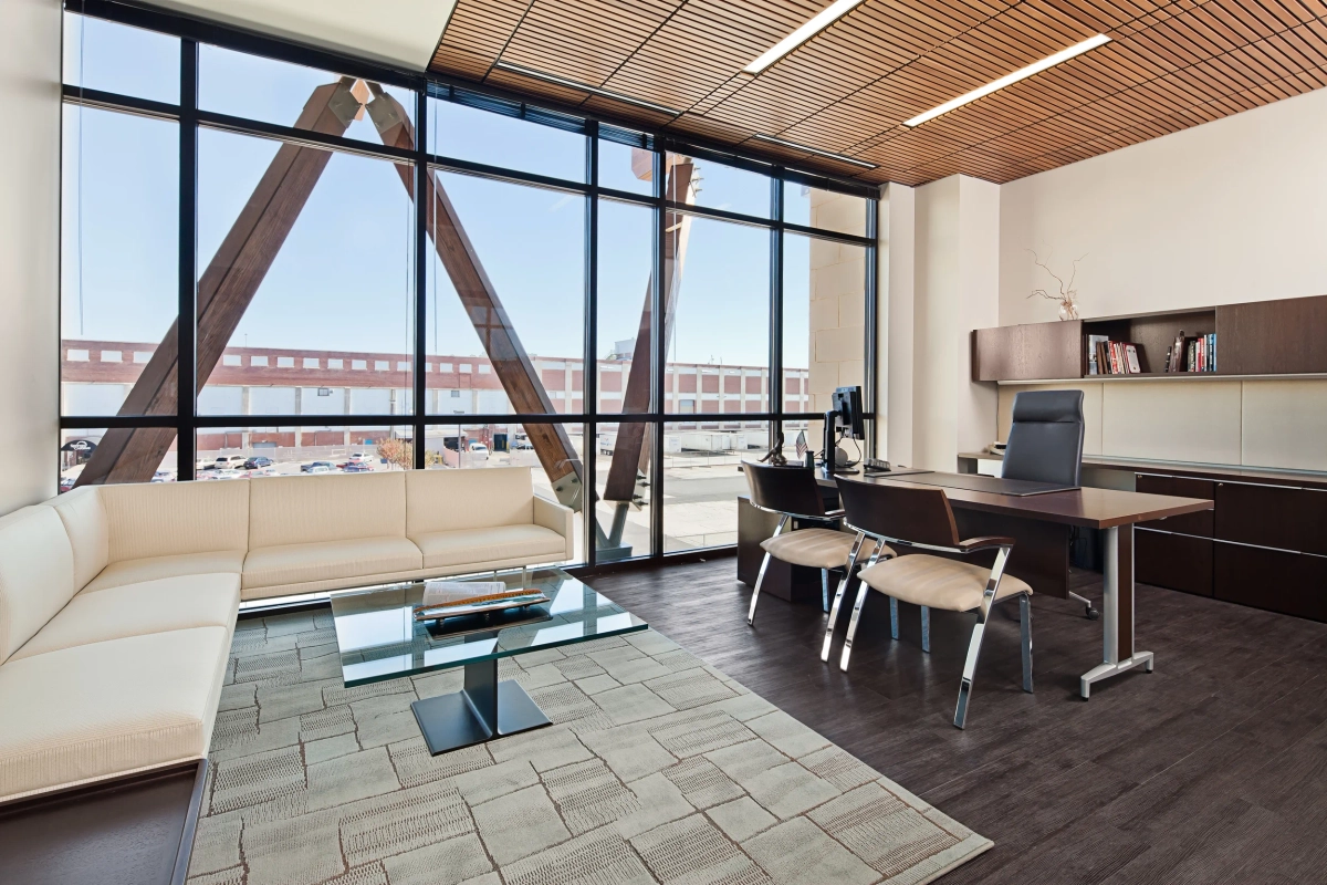 Corner executive office with floor-to-ceiling windows, exposed structural wood A-frame beam, cream L-shaped sofa, glass coffee table, dark wood desk with ergonomic chair, and warm wood ceiling slats
