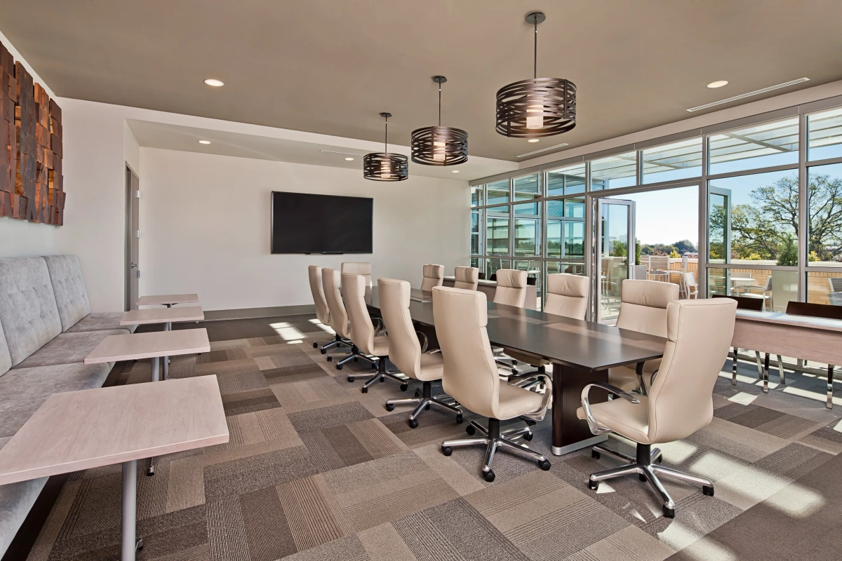 Large corporate boardroom with a long dark conference table, cream leather executive chairs, three bronze drum pendant lights, wall-mounted display screen, geometric wood accent wall panel, and full-height windows