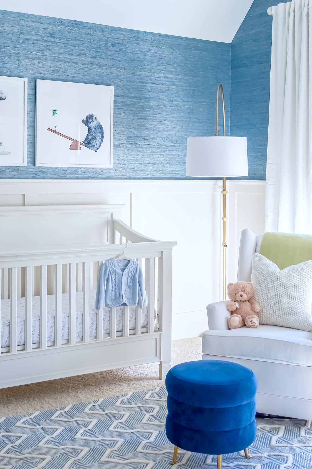 Boy nursery corner with a white crib, blue cable-knit sweater hanging, whimsical animal art prints, blue grasscloth walls, white glider with teddy bear, and navy velvet ottoman