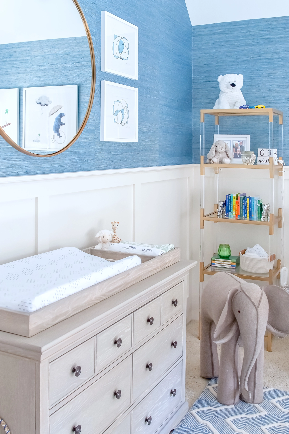 Boy nursery detail with a light wood dresser and changing pad, brass round mirror, blue grasscloth wallpaper, framed art prints, acrylic and brass bookshelf with toys and books