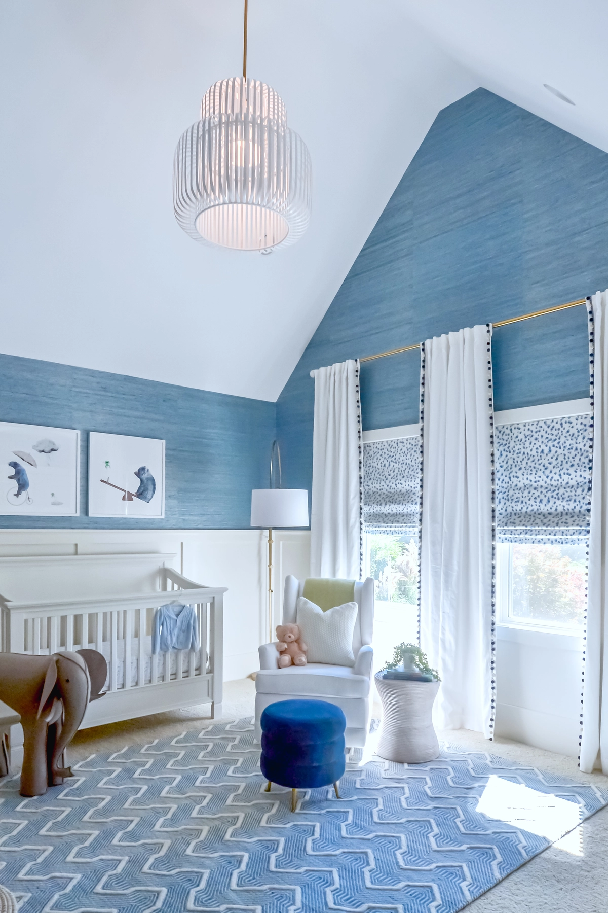 Boy nursery with blue grasscloth wallpaper, vaulted ceiling, white crib, white glider chair, navy velvet ottoman, slatted pendant light, pom-pom curtains, and blue chevron rug