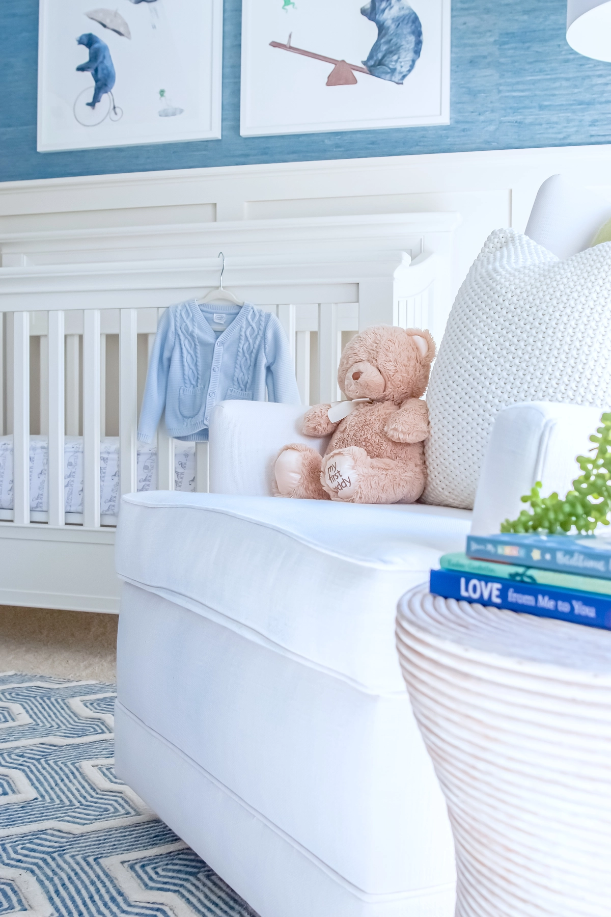 Close-up of a white glider chair with a teddy bear, knit pillow, white crib with animal prints above, blue grasscloth wallpaper, and a woven side table with stacked books