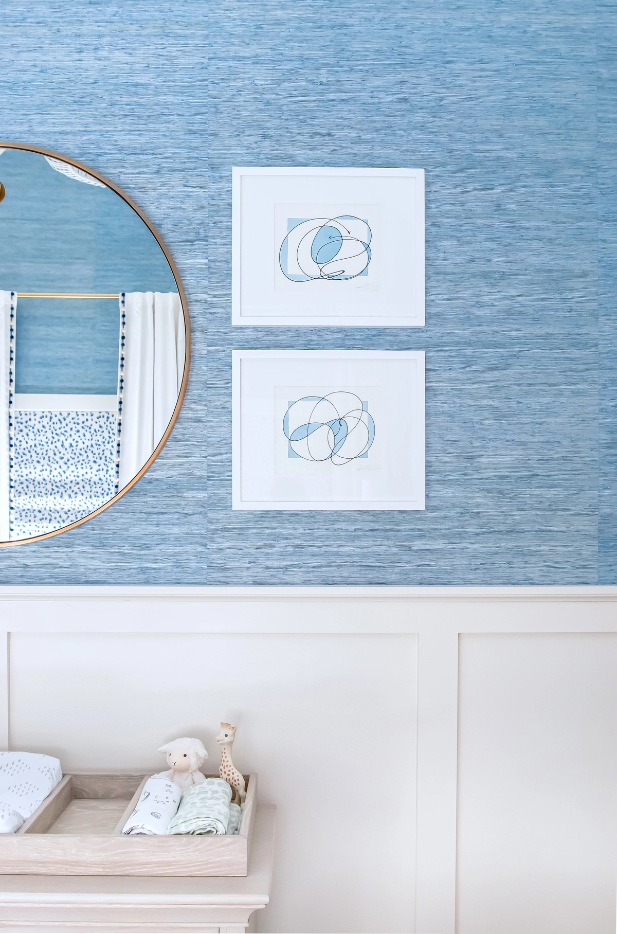 Close-up of blue grasscloth wallpaper with brass round mirror, abstract line art prints, and a light wood dresser with changing pad and stuffed toys