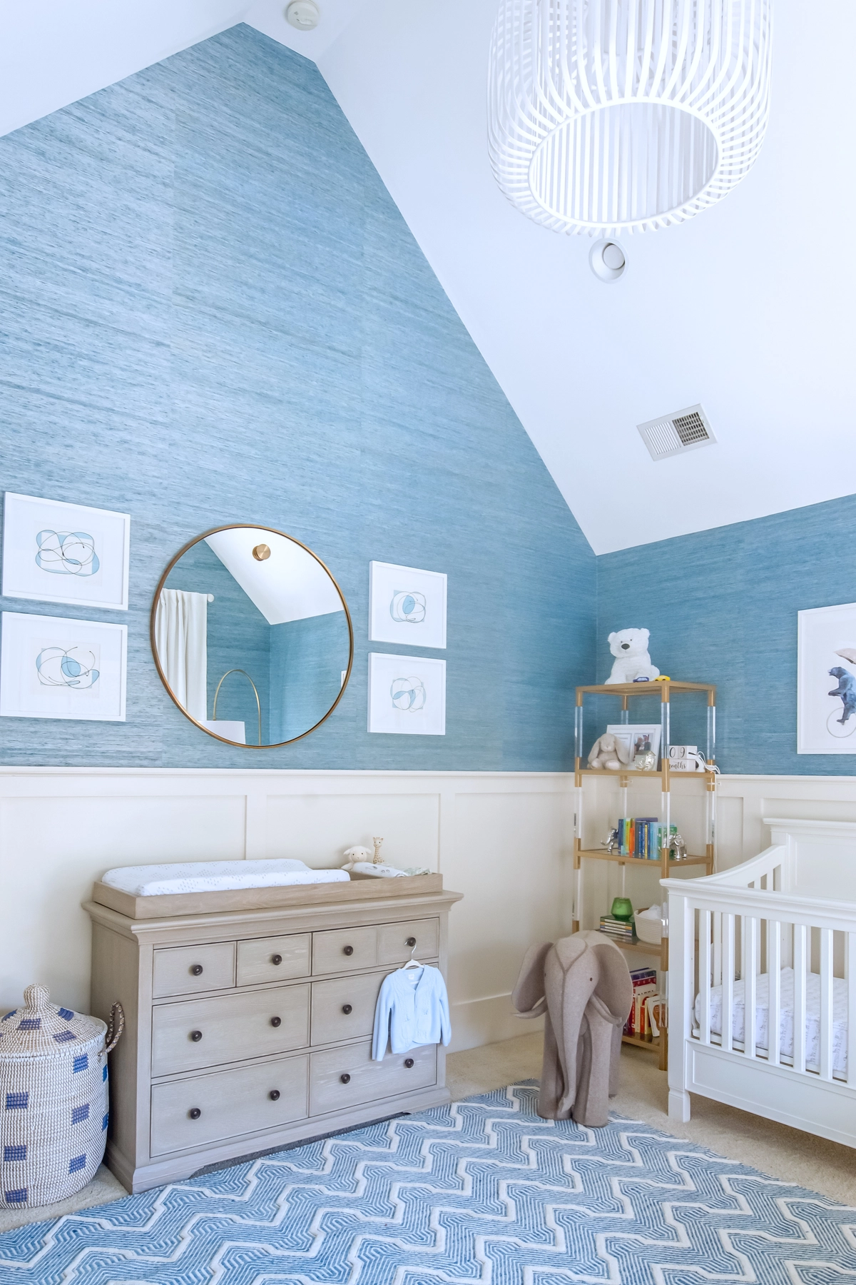Wide view of a boy nursery with blue grasscloth wallpaper, white wainscoting, round brass mirror, light wood dresser, white crib, stuffed elephant, and blue chevron rug